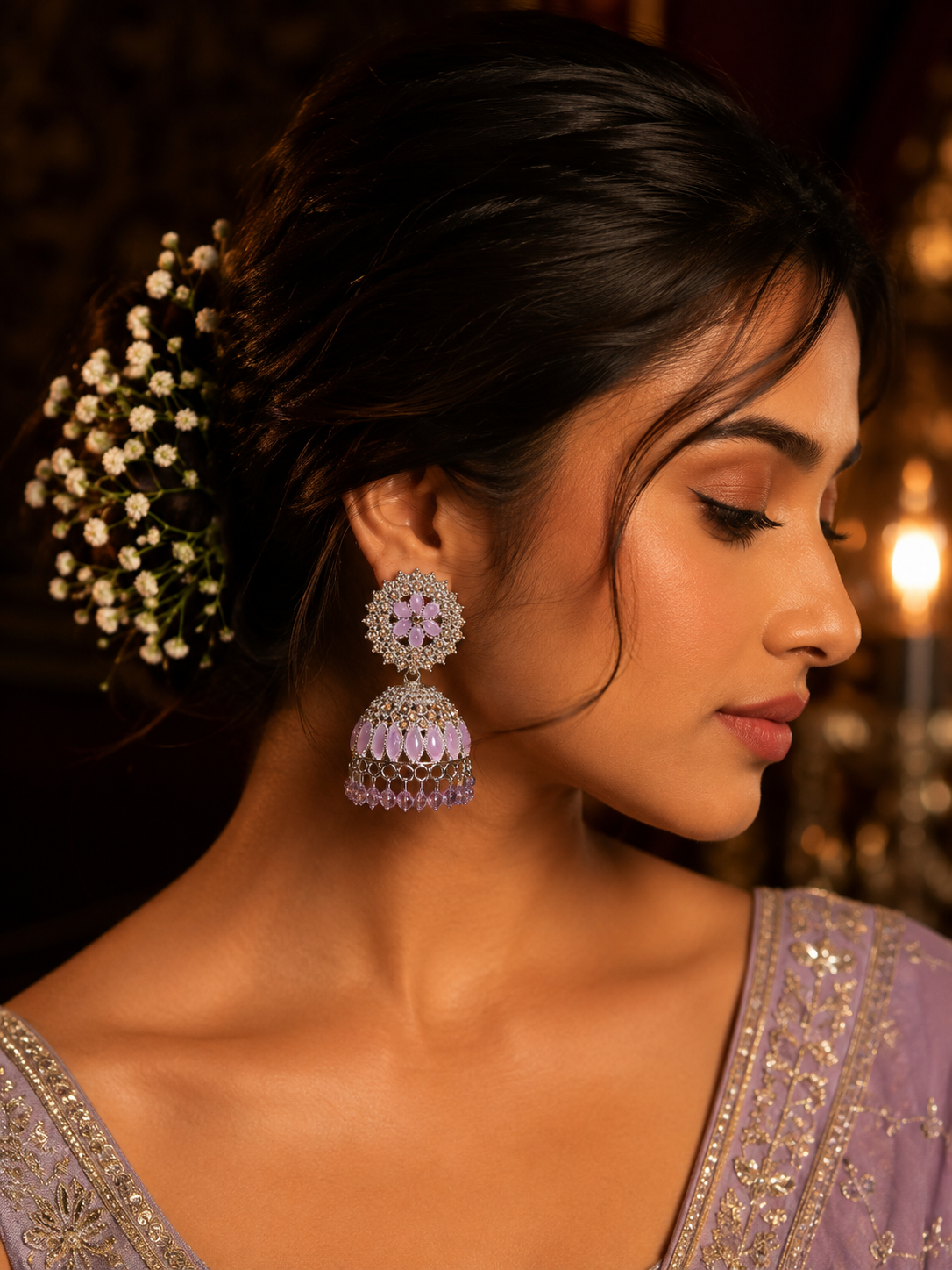 A close-up side profile of a woman wearing ornate silver-toned jhumka earrings with light purple stone accents, styled with a floral hair accessory and a lavender embroidered garment.
