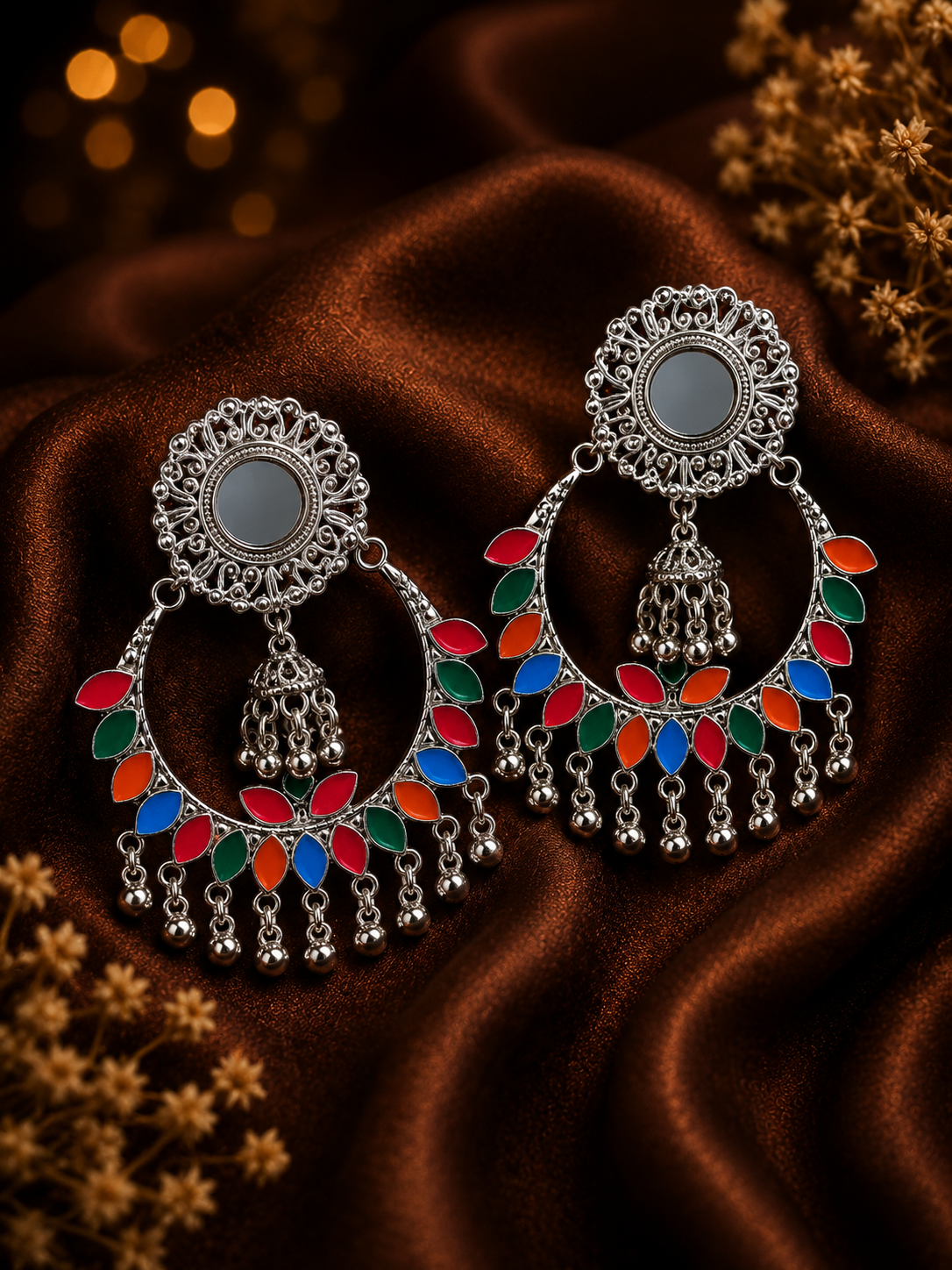 A pair of ornate silver-toned Indian chandelier earrings with colorful enamel details in red, blue, and green, displayed on a draped brown satin fabric background.