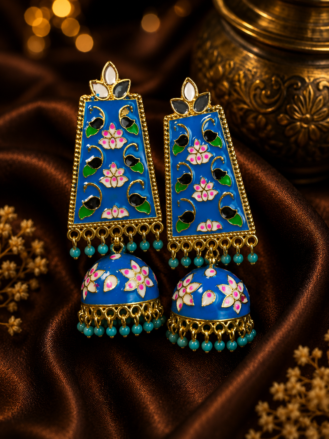 A pair of ornate Indian jhumka earrings with vibrant blue enamel, pink lotus floral motifs, and green peacock details, set in gold with turquoise bead fringe, displayed on a brown satin fabric.