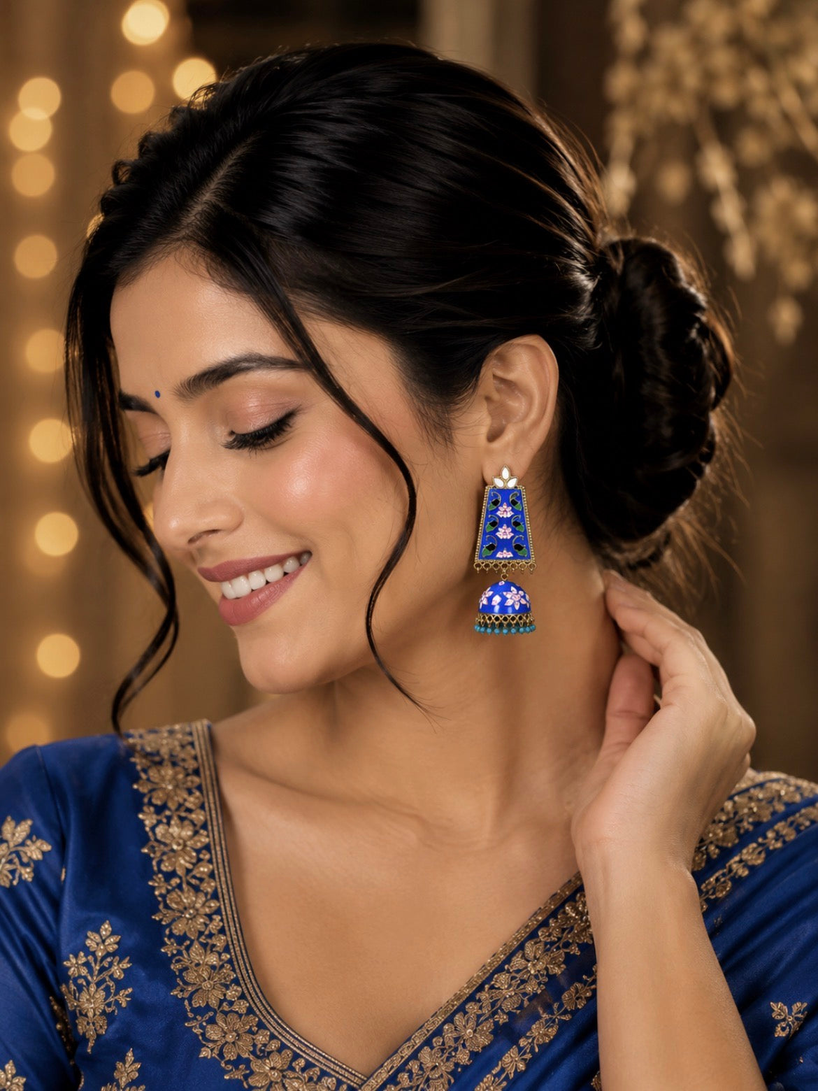 A woman wearing a blue embroidered traditional outfit and a matching blue jhumka earring with gold and white embellishments, set against a warm, softly blurred background with bokeh lights.