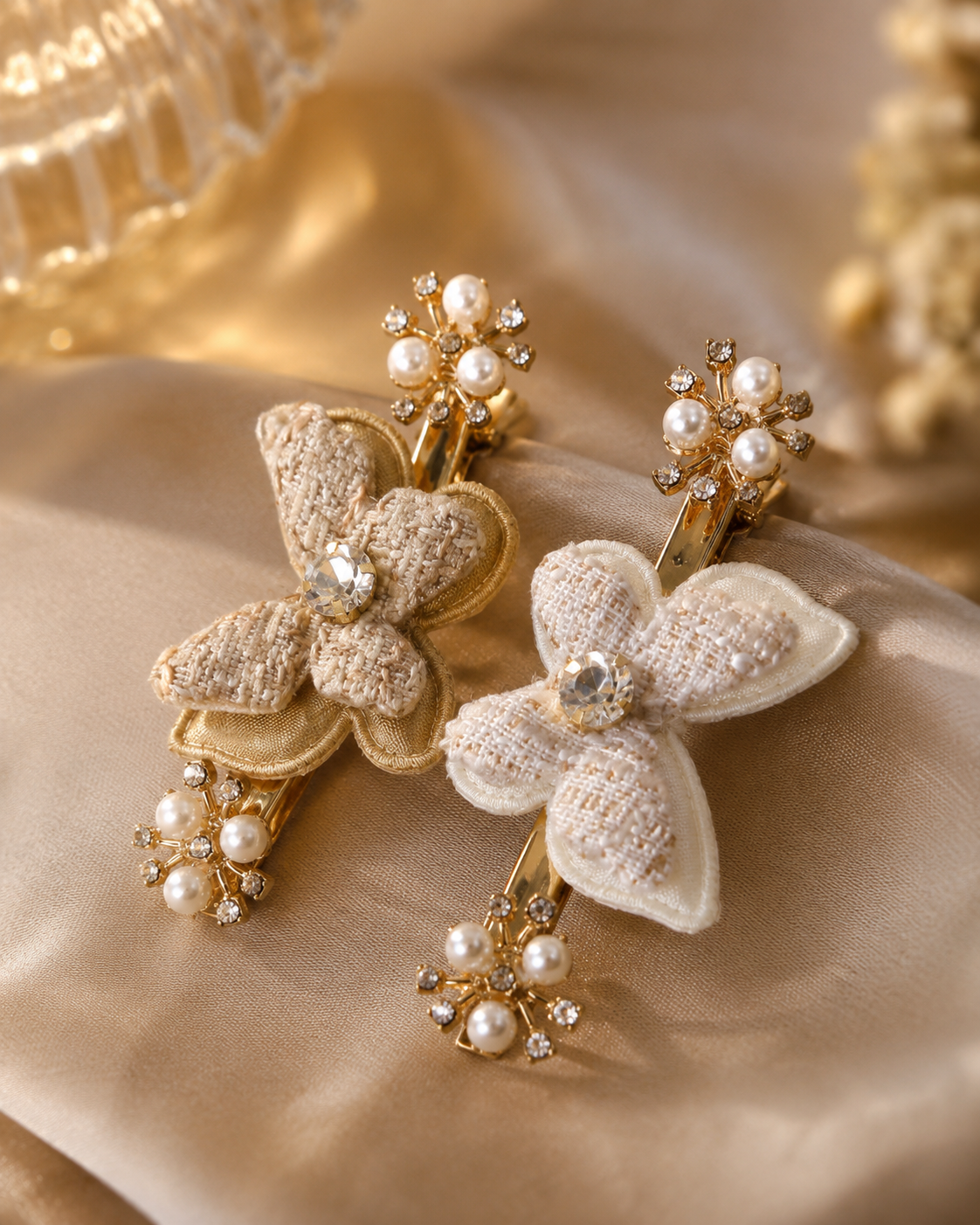 Two decorative hair claw clips styled with embroidered butterfly motifs, pearl beads, and rhinestone accents on a draped beige satin fabric background.