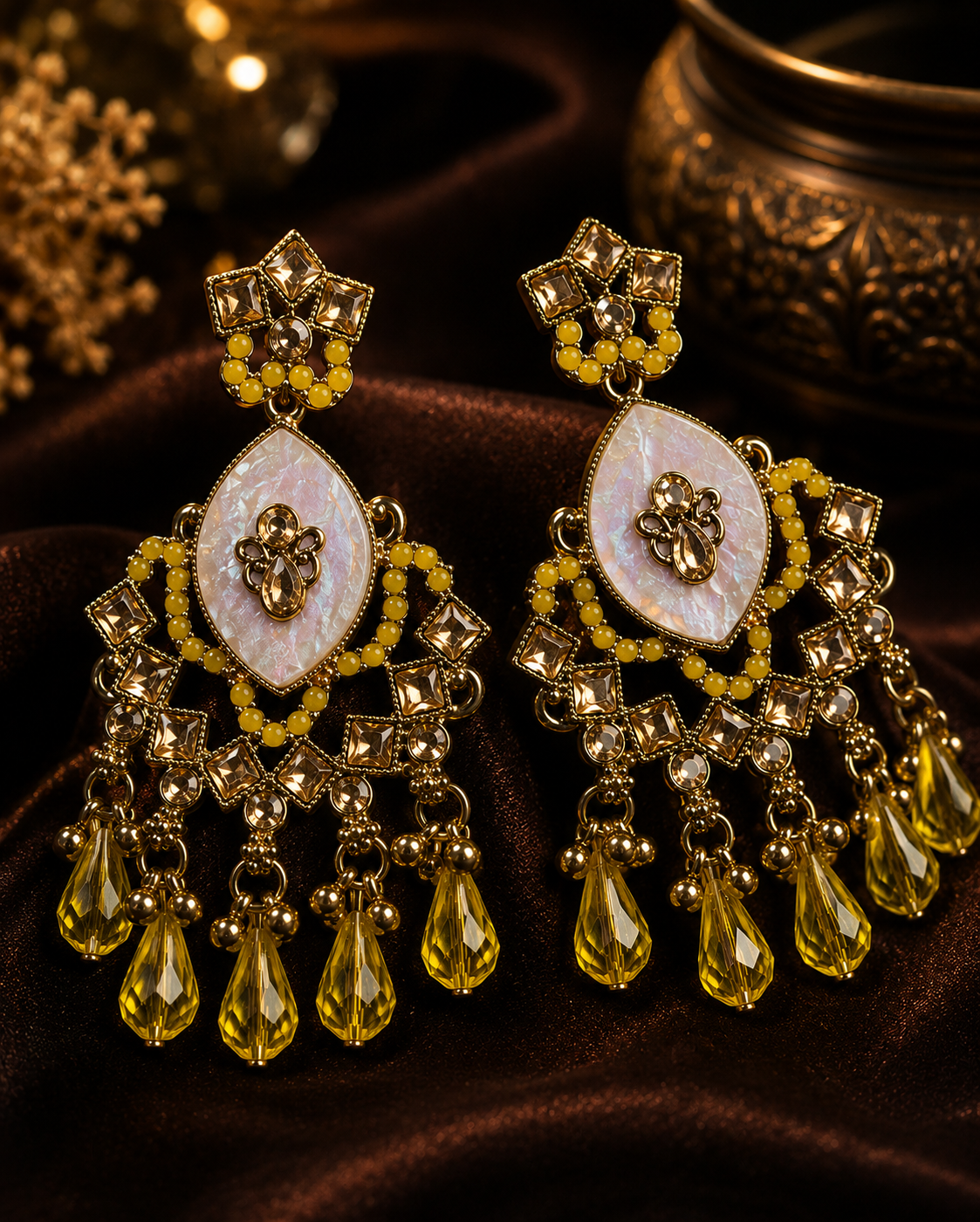 A pair of ornate Indian-style gold-toned dangle earrings with iridescent white mother-of-pearl teardrop centers, yellow beaded details, and dangling yellow crystal drops, displayed on dark brown satin fabric.