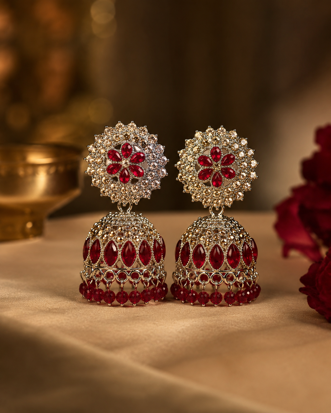 A pair of ornate Indian jhumka earrings with red gemstone floral motifs and teardrop-shaped red stones, set in silver-toned metal with clear stones, displayed on a beige fabric surface.
