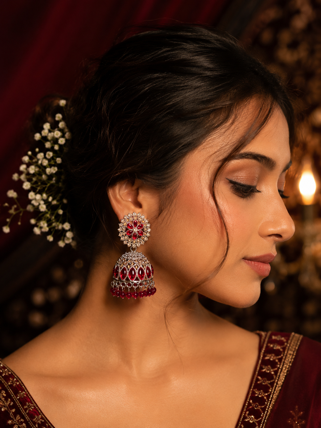 A close-up side profile of a woman wearing a traditional Indian jhumka earring. The earring features a silver-toned filigree body with deep red stones and a dangling red beaded bottom.