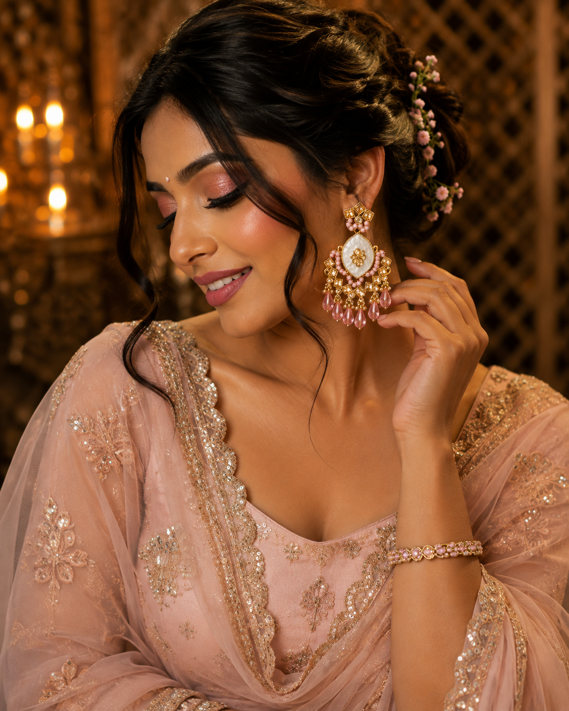 A woman wearing a light pink embellished traditional outfit and matching jewelry, including a large pink and gold statement earring and a bracelet, posed against a warm, softly lit background.