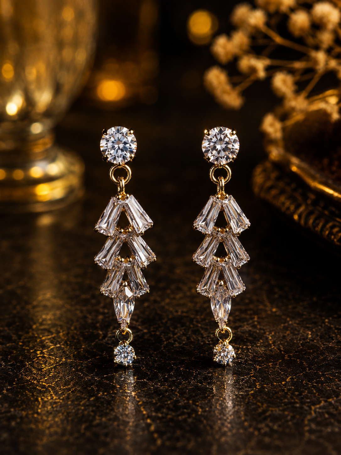 A pair of gold drop earrings with clear faceted gemstones, displayed on a dark reflective surface with warm golden bokeh in the background.