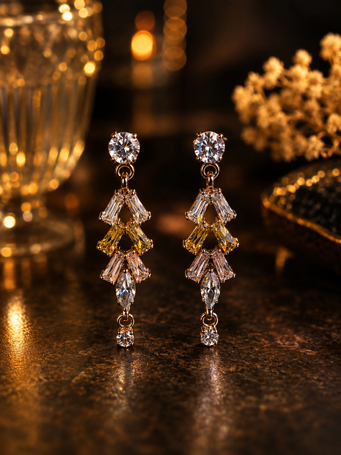 A pair of gold drop earrings with clear and yellow gemstones, displayed on a dark reflective surface with warm bokeh background lights.