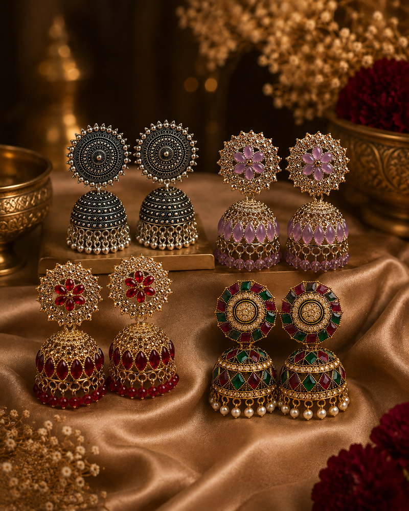 Four pairs of ornate Indian jhumka earrings arranged on a draped satin fabric. The earrings feature intricate gold-plated metalwork, beadwork, and colorful stone inlays in red, green, purple, and black.