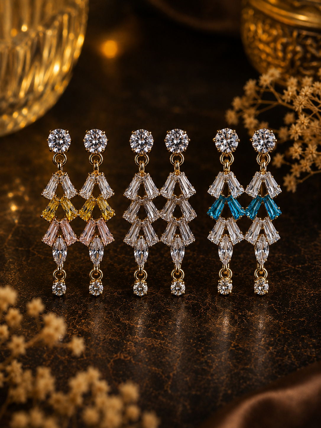 Six pairs of gold-tone drop earrings with clear faceted crystals and colored crystal accents in yellow, pink, and blue, arranged on a dark textured surface with decorative dried flowers and warm golden background elements.