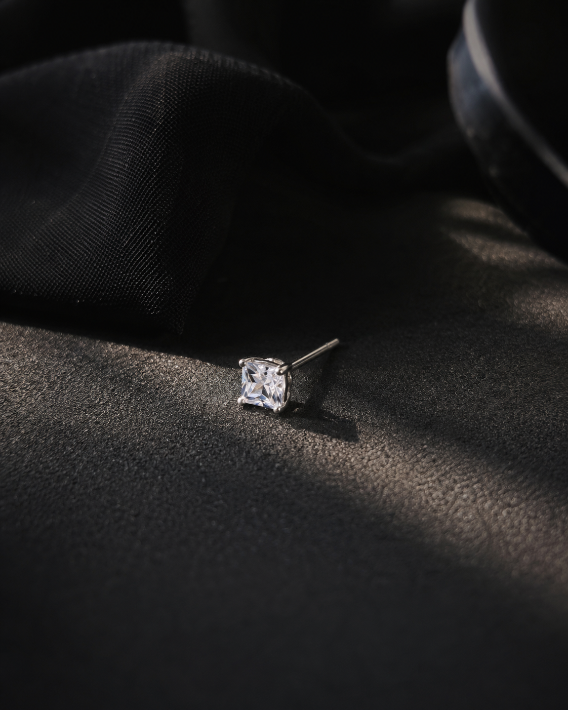 A single solitaire ring with a clear princess-cut gemstone in a silver setting, resting on a dark textured surface with dramatic shadow lighting.