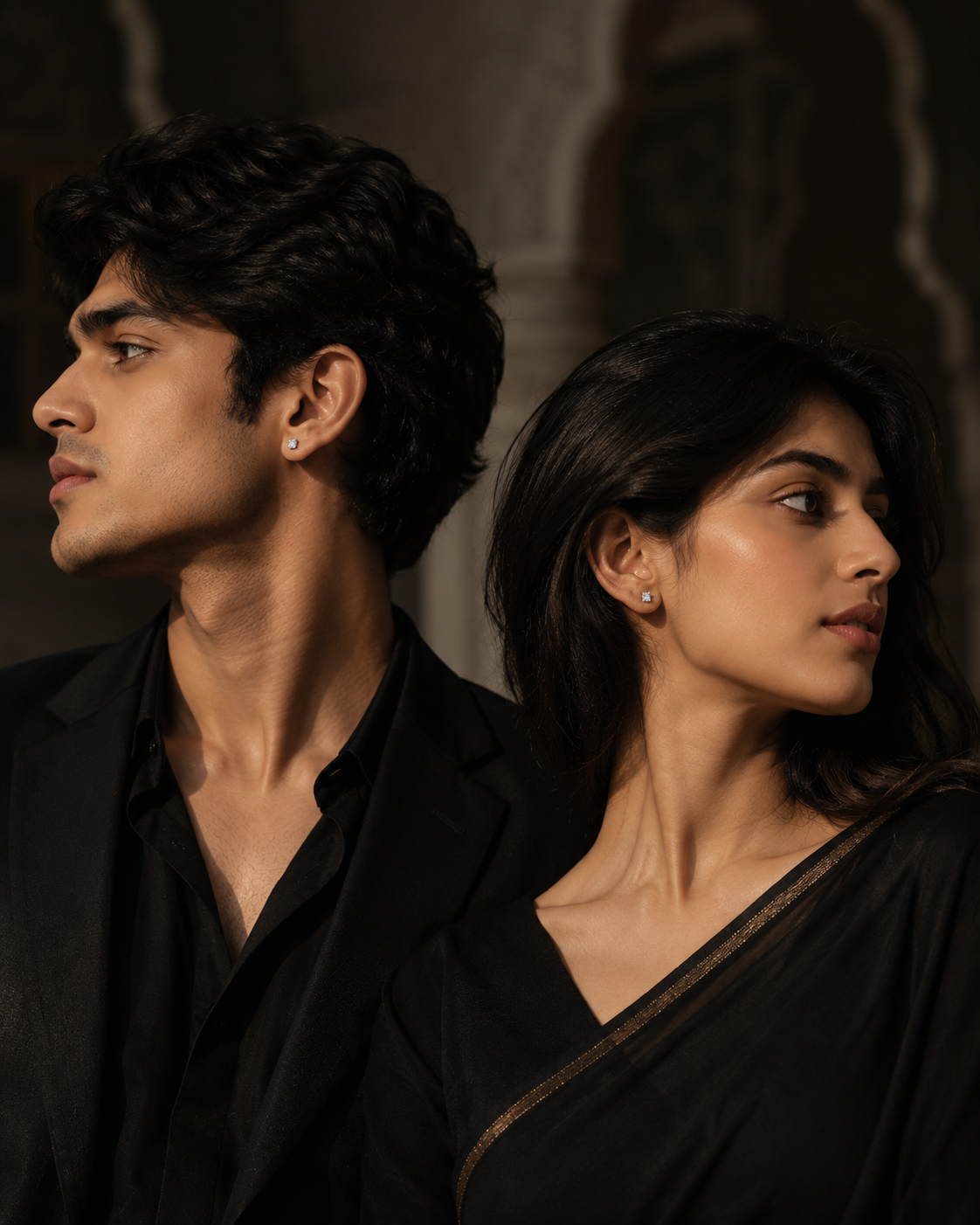 A man and a woman in black clothing posed in profile against a dark, softly lit architectural background. The man wears a black shirt and jacket, and the woman wears a black sari with a subtle gold-toned border.