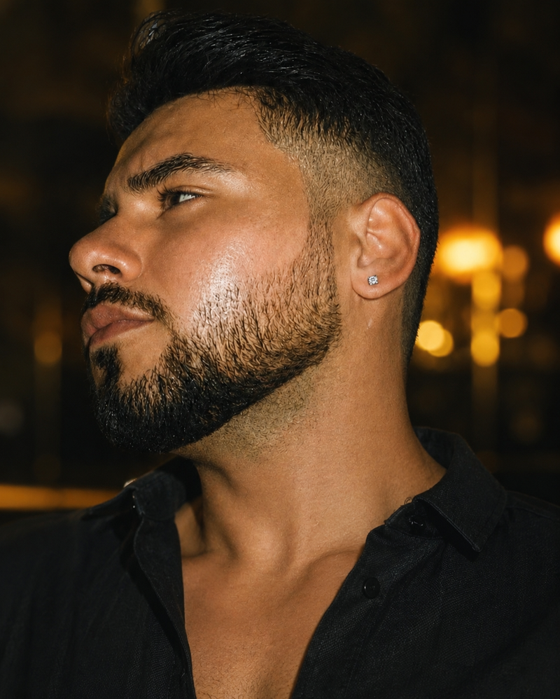 A close-up profile portrait of a man with a short beard and styled dark hair, wearing a small silver stud earring and a black button-up shirt, set against a warm, blurred bokeh background.